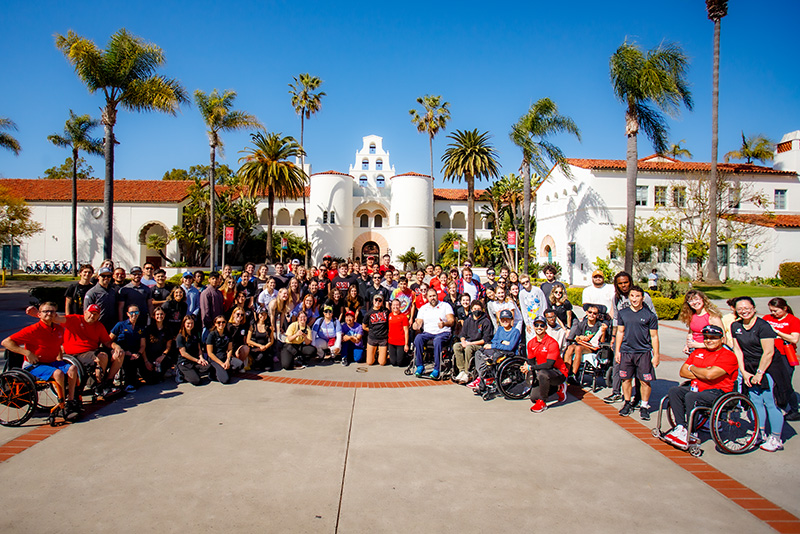 Adapted Athletics athletes and supporting team members posing together in front of Hepner Hall, San Diego State University.