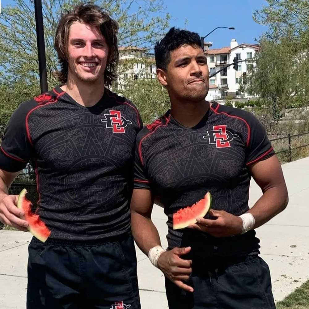 Two smiling men's rugby club members eating watermelon after the game