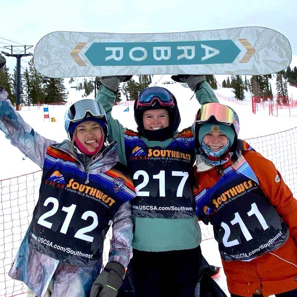 Three smiling ski and snowboard club members at the snowy mountain, center member holding up snowboard above his head