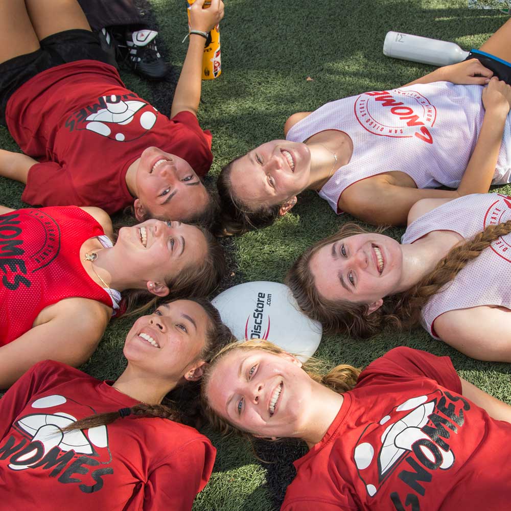 Six smiling women's ultimate frisbee members laying down on grass with frisbee disk in the center