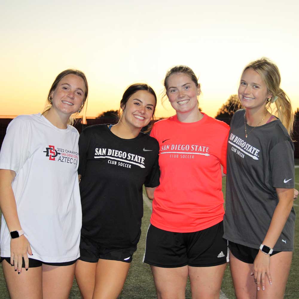 Four smiling women's soccer club members on the field at sunset