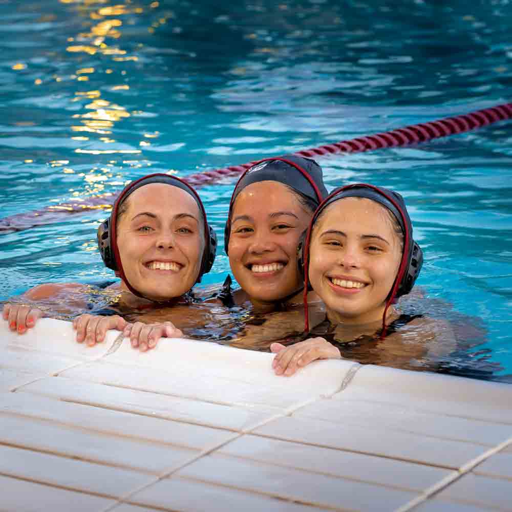 Three smiling SDSU women's water polo players at the edge of the pool